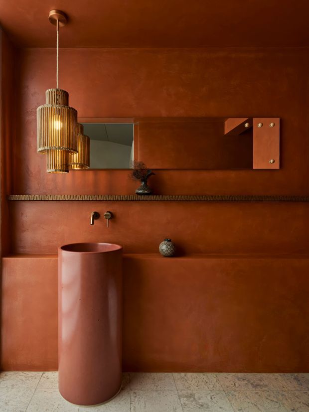 A richly monochromatic terracotta bathroom featuring a cylindrical freestanding basin, wall-mounted brass-toned tapware, a long timber-framed mirror, integrated wall ledges, and a sculptural ribbed pendant light. The walls and built-in surfaces have a smooth, mottled plaster finish, while the floor is clad in pale textured stone or terrazzo-like tiles. The atmosphere is warm, moody, refined, and highly sculptural.