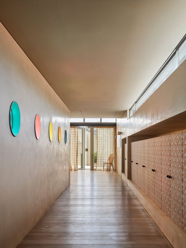 A long, narrow interior hallway or entry passage with warm, softly textured plaster walls and dark timber flooring. The left wall features a series of colorful circular decorative insets, while the right side contains extensive built-in joinery or shelving with a patterned tile backsplash and small dark fixtures. A high clerestory window runs along the right side, and glazed doors at the far end bring in natural light. The space has a carefully designed, retro-influenced contemporary character.