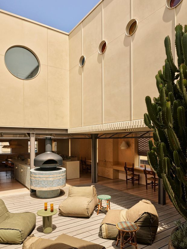 A sunlit contemporary courtyard centered on a raised timber deck, with a rounded tiled outdoor pizza oven, low beanbag seating, small stools, and a dining area beneath a covered overhang. Tall warm beige rendered wall panels feature distinctive circular porthole-style windows, while a large sculptural cactus and other planting occupy the right side. The space has a warm, relaxed indoor-outdoor character with strong geometric architectural detailing.