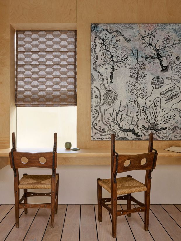 A warm, wood-lined interior study or dining/work area with two vintage wooden chairs positioned at a long built-in desk or console. The room features pale timber wall panelling and wide timber floorboards, a geometric woven blind over a window, and a large botanical and abstract artwork mounted above the desk. Small decorative objects sit on the built-in surface. The composition has a handcrafted, calm, earthy character.