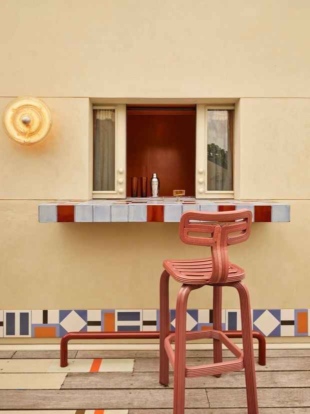 A stylised architectural exterior vignette featuring a compact wall-mounted bar or serving counter beneath a recessed window. The warm beige rendered wall has a colourful geometric tiled band near the base, while the counter uses alternating pale blue-grey and terracotta-red tiles. A sculptural terracotta-red bar stool sits on a weathered timber deck, with bottles and glassware displayed in the recessed opening. The composition has a playful, highly graphic character with strong geometric detailing and warm afternoon light.