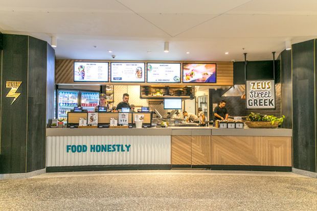 Front-facing view of a contemporary Greek street-food takeaway counter in a commercial interior. The service area features illuminated menu boards, refrigerated drink displays, food preparation equipment, a dark green and timber-toned counter facade, ribbed white paneling, and prominent brand signage. The space has bright overhead lighting and a clean, functional hospitality fit-out.