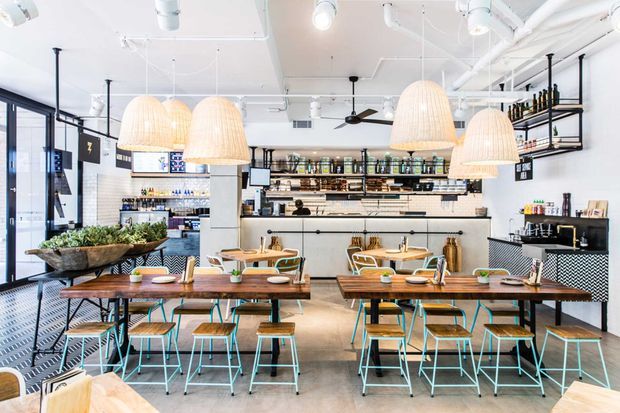 Bright contemporary café interior with communal timber dining tables, turquoise metal stools and chairs, woven pendant lights, a long service counter, open shelving stocked with bottles and products, and exposed white ceiling services. Black-framed glazing and patterned tile detailing add industrial character.