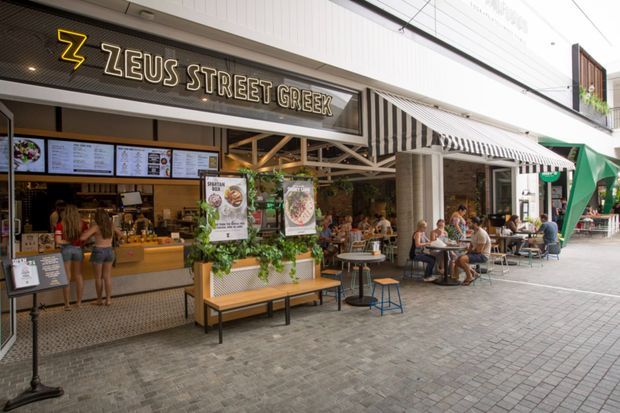Street-facing Greek food venue in a covered commercial laneway, with a prominent Zeus Street Greek sign, open service counter, menu boards, striped awnings, outdoor café tables, and bench seating. The space has a lively casual dining atmosphere with exposed structural framing, tiled paving, and extensive shopfront glazing.