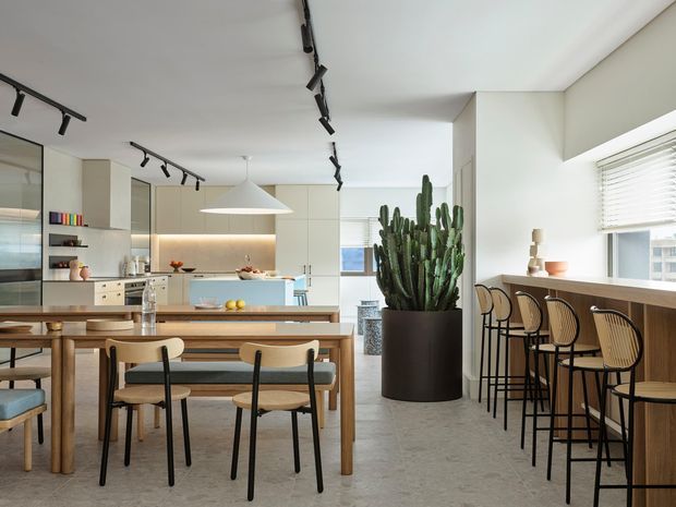 Bright open-plan kitchen and dining area with a long timber dining table, upholstered dining chairs, pale built-in cabinetry, a kitchen island, and a built-in window-side breakfast bar with stools. Black track lighting, a large sculptural pendant, concrete-look flooring, and a prominent potted cactus add contemporary character.