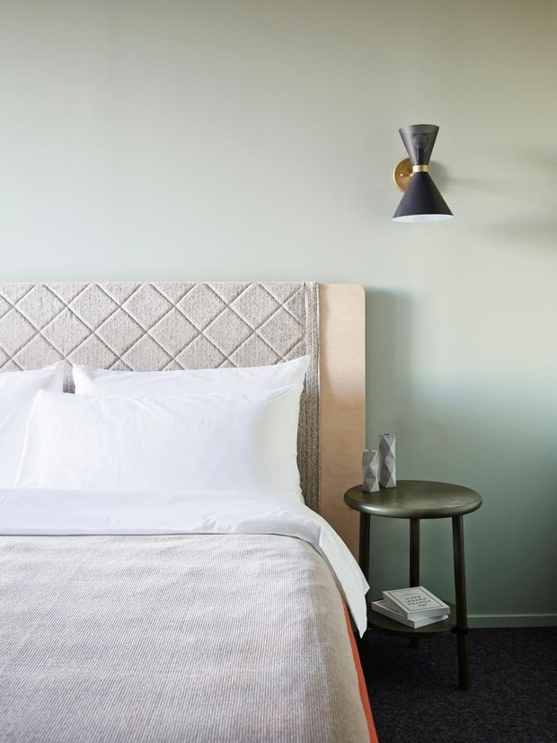 A softly styled bedroom corner featuring a bed with a pale upholstered diamond-quilted headboard, white bedding, and a light timber side panel. A dark round bedside table holds decorative objects and books, while a sculptural black-and-brass wall sconce is mounted on a pale sage green wall. Dark carpet grounds the room.