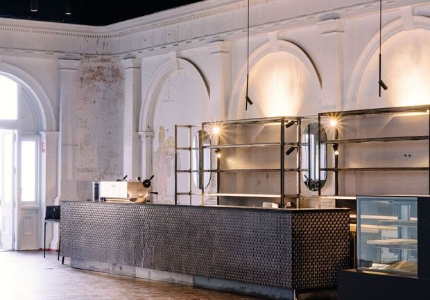 A refined commercial café or bar interior in a repurposed historic room, featuring ornate white arched wall mouldings, a weathered plaster finish, polished timber flooring, and a long dark metal-clad service counter. Open metal-framed shelving with integrated warm lighting sits behind the counter, alongside a glass pastry display case. Tall arched windows and elaborate classical detailing contrast with the contemporary industrial fit-out.