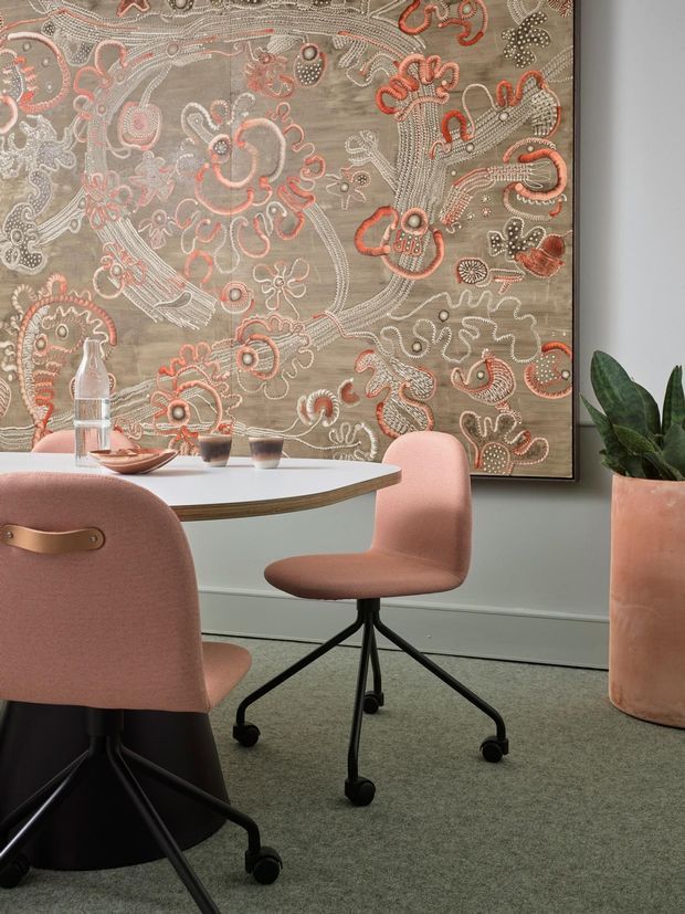A styled dining area with a white oval table, blush pink upholstered swivel chairs on black bases, and a large framed abstract textile artwork featuring intricate coral, cream, and grey patterns. A terracotta planter with a leafy indoor plant sits to the right, while neutral grey-green walls and carpet create a muted, contemporary setting.