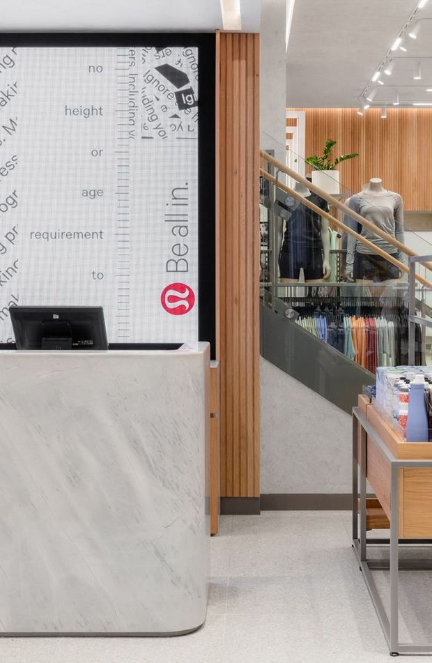 A bright contemporary retail clothing store interior with a curved white marble reception counter in the foreground, a large branded graphic backdrop, timber slat wall cladding, glass balustrades, clothing displays, mannequins, and a stairway leading to a mezzanine or upper level. The space has polished light flooring and linear track lighting.