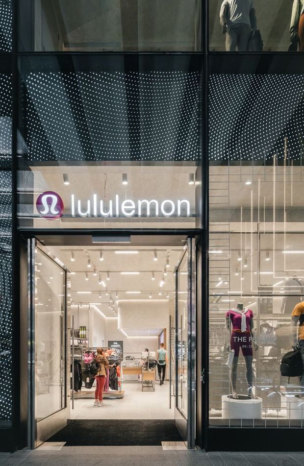 A street-level view into a Lululemon retail store through a tall glazed entrance. The storefront has dark metal framing, expansive glass, a perforated dark facade screen creating a dotted pattern, bright minimalist retail lighting, pale interior finishes, clothing displays and mannequins. The image is primarily an exterior commercial storefront with the shop interior visible through the glazing.