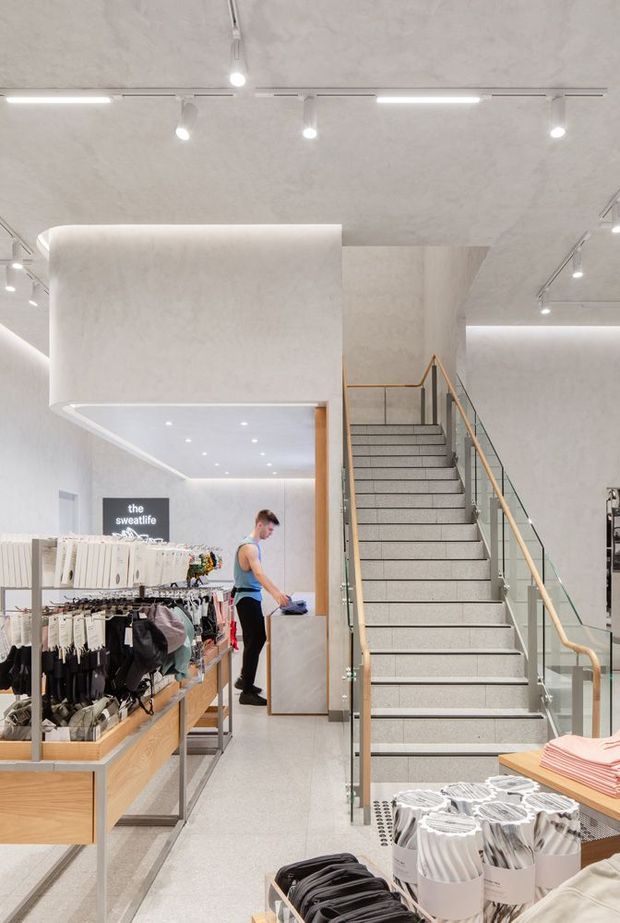 Bright contemporary retail clothing store interior with pale textured walls and ceiling, integrated linear and track lighting, timber display tables, clothing racks, a central staircase with glass balustrades and brass-toned handrails, and a mezzanine or upper-level circulation area. The space has a clean, refined commercial fit-out with predominantly light neutral finishes.