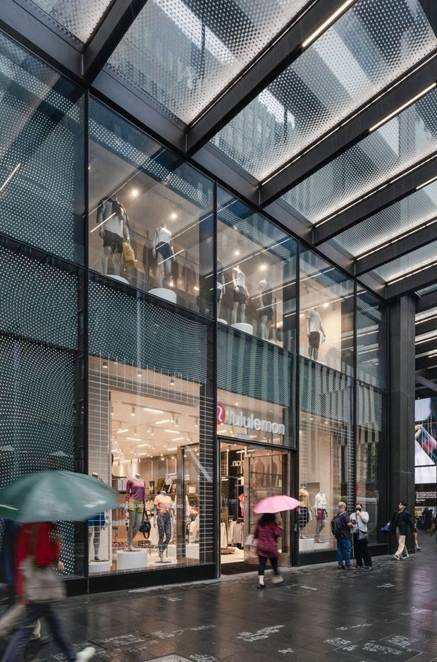 Street-level view of a contemporary multi-storey retail storefront, featuring extensive floor-to-ceiling glazing, black structural framing, perforated metal facade panels, illuminated linear ceiling elements, and mannequins displayed inside a Lululemon shop. Pedestrians with umbrellas occupy the wet urban walkway in front.