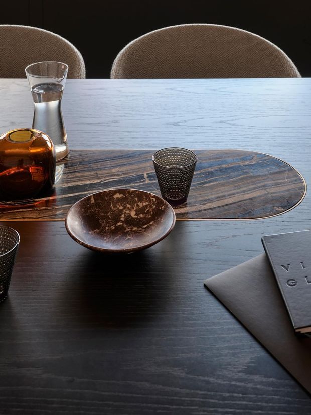 Close-up still-life view of a dark timber dining table with a rounded rectangular natural-stone or marble-look placemat, sculptural ceramic and glass vessels, textured drinking glasses, and two upholstered dining chairs in the background. The composition is refined and understated, with warm brown accents against a deep charcoal-brown table surface.