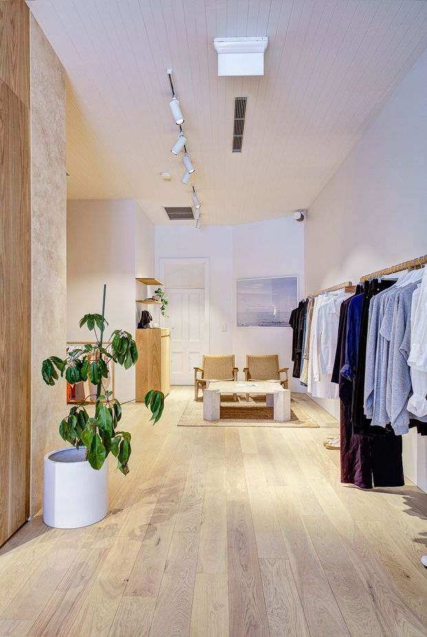 A bright, narrow contemporary clothing boutique with pale timber flooring and a matching slatted timber ceiling. Clothing is displayed on an open rail along the right wall, while the left side features timber joinery, a full-height textured panel, shelving, and a large potted plant. At the rear is a small seating area with woven chairs, a low coffee table, and a neutral rug. Track lighting and linear ceiling vents provide functional illumination and ventilation.