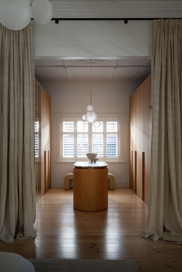 A refined, narrow dressing room or walk-in wardrobe framed by full-height neutral linen curtains. Floor-to-ceiling timber cabinetry lines both sides, with a central cylindrical timber island featuring a pale stone top. A built-in bench sits beneath shuttered windows at the far end, while layered decorative plaster ceiling detailing and clustered spherical pendant lights add a classic architectural character. The space combines warm timber, soft beige textiles, white joinery, and natural daylight.