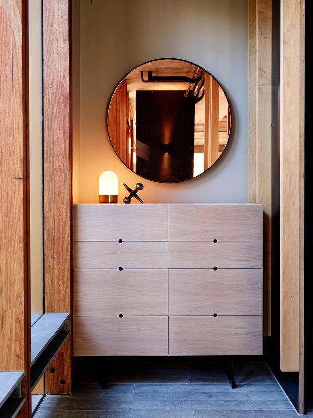A compact, warmly lit entryway or dressing area featuring a pale timber six-drawer dresser, round wall mirror, sculptural black table lamp, and extensive vertical timber wall framing. Dark tiled flooring and black accents create a refined contemporary look, while the mirror reflects exposed services and timber detailing beyond.