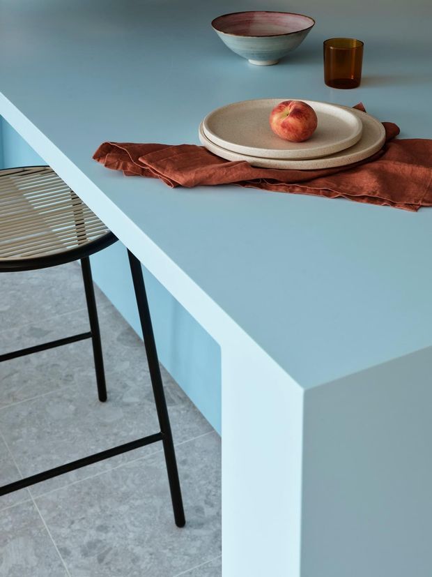 Close-up interior styling photograph of a pale blue dining table with a thick squared edge, paired with a black metal-framed woven-seat chair. A rust-red linen runner, ceramic plates and bowl, amber glass tumbler, and peach provide a warm tabletop arrangement. The space has cool grey tiled flooring and a refined contemporary palette.