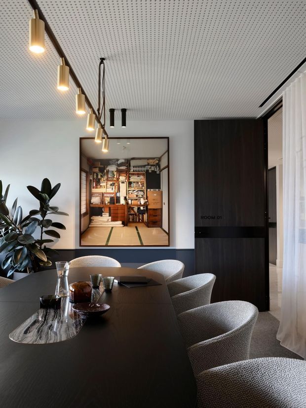 A refined contemporary dining room with a long dark dining table, sculptural upholstered dining chairs, decorative tableware, and linear pendant lighting. The room features a perforated white ceiling, pale walls, dark timber or black built-in joinery, sheer curtains, and a large framed opening or artwork revealing a compact timber-lined study or library beyond. A leafy indoor plant adds a natural accent.