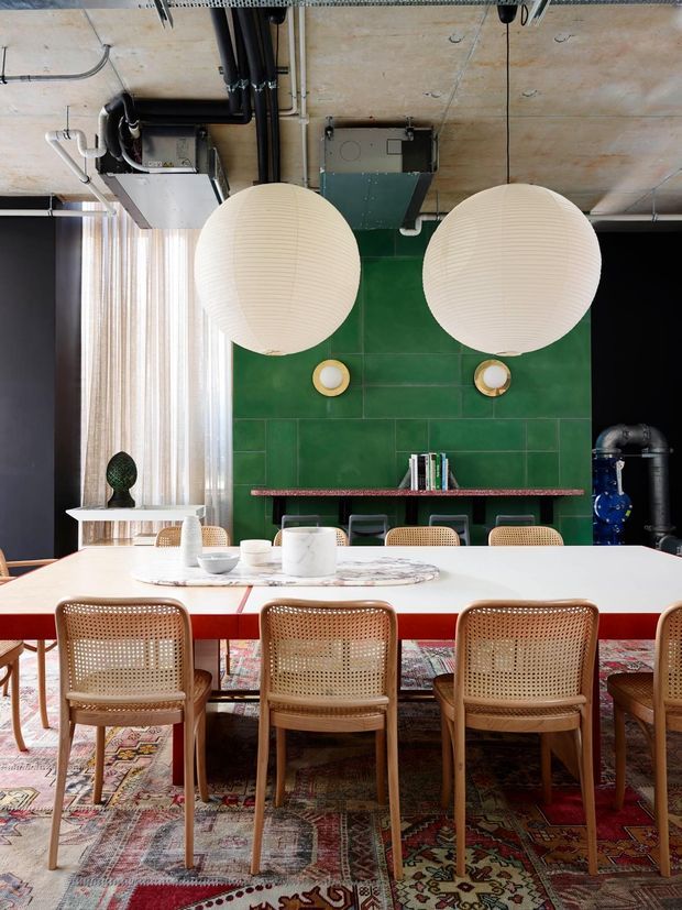 A stylish dining area with a large white rectangular dining table surrounded by woven cane-back chairs. The room features a bold green tiled feature wall with a narrow red shelf, oversized spherical pendant lights, exposed concrete ceiling services and ductwork, dark wall panelling, sheer curtains, and a richly patterned traditional rug. The overall scheme combines industrial architectural elements with colourful contemporary and vintage-inspired furnishings.