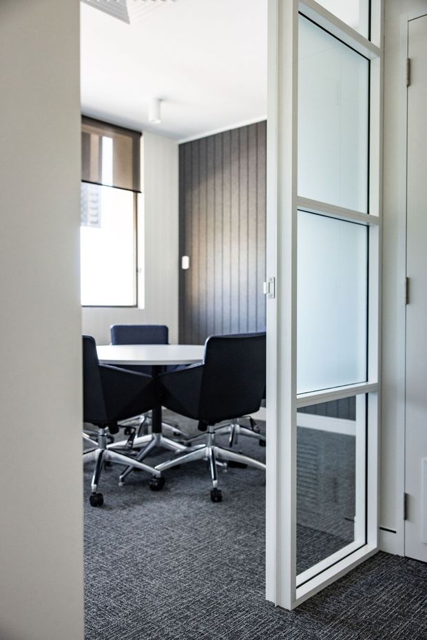 A contemporary commercial meeting room viewed through an open glazed door. The room contains a white conference table, several black swivel office chairs, grey carpet, a dark vertically panelled feature wall, and a tall window with a roller blind. White walls, ceiling, and framed glass doors create a clean, professional atmosphere.