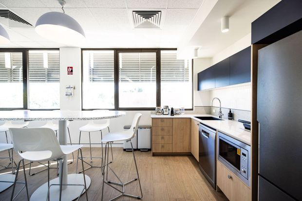 Bright contemporary kitchenette and break-out dining area with timber-look flooring, wood-grain base cabinetry, dark navy overhead cabinetry and tall storage, white benchtops, integrated appliances, and a central high table with white bar stools. Large windows with horizontal blinds provide abundant natural light, while exposed acoustic ceiling tiles and suspended pendant lighting suggest a commercial office setting.