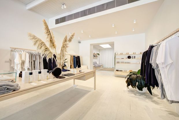 Bright, minimalist clothing boutique with pale timber flooring, cream walls, open clothing rails, built-in display shelving, a long central timber display table, recessed spotlights, and a decorative dried-grass arrangement. The space has a clean, airy retail fit-out with neutral garments and product displays.