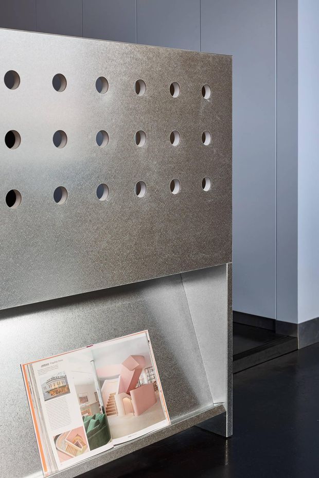 A close-up of a modern architectural display or presentation stand in a minimalist interior. The freestanding metallic panel has a textured silver-grey finish and a regular grid of circular cut-outs, with a slanted lower shelf holding an open architecture or interiors magazine. Dark flooring and tall grey built-in wall panels create a restrained, contemporary exhibition-like setting.