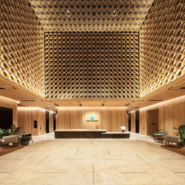 A grand contemporary commercial lobby or reception area with a central reception desk, extensive warm timber wall panelling, pale stone flooring, and lounge seating with indoor plants. The defining feature is a dramatic recessed ceiling installation composed of hundreds of circular timber or wood-toned apertures, creating a textured geometric canopy above the reception zone. BNP Paribas signage is visible on the rear wall.