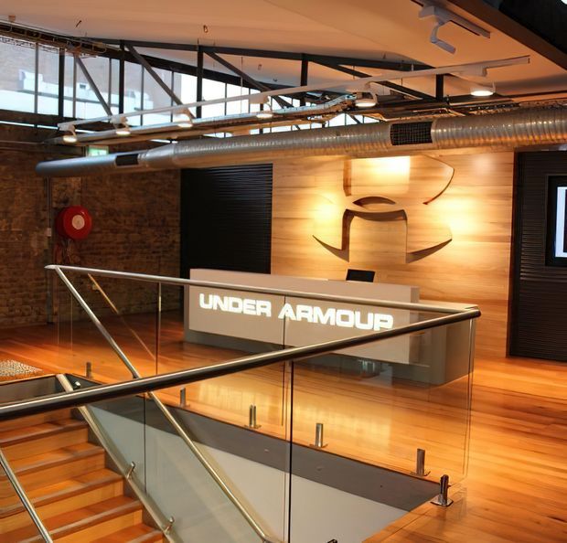 Interior view of a branded commercial reception or showroom space with a prominent illuminated 'Under Armour' sign, exposed industrial services and ductwork, timber wall and flooring, exposed brick, black-framed glazing, and a glass-and-metal stair balustrade.