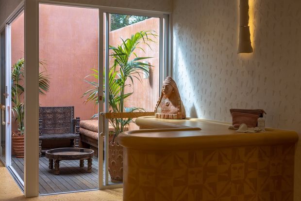 Warmly lit interior featuring a curved ochre reception or bar counter with patterned tile detailing, a textured light wall, sculptural pendant light, and a carved decorative figure. Full-height glazed doors open to a small timber-decked courtyard or terrace furnished with seating and tropical plants.