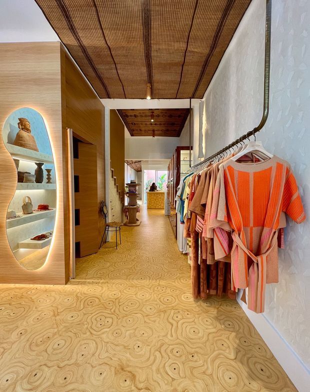 A warmly lit boutique retail clothing store with a long hallway-like layout. Clothing is displayed on a wall-mounted rail at right, while illuminated arched display niches with accessories are built into timber joinery on the left. The space features highly patterned light timber flooring, timber wall panels, white textured walls, and a woven timber or rattan ceiling treatment, creating a playful, crafted, organic interior.