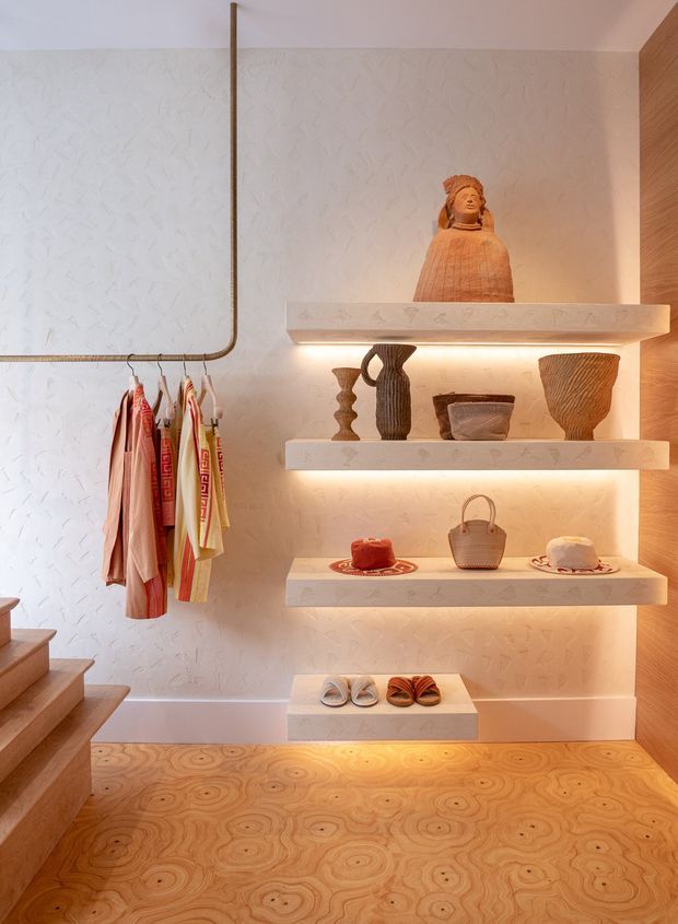 Warmly lit boutique-style retail display or dressing area with floating illuminated shelves holding sculptural ceramics, textiles, bags, hats and shoes. A textured off-white plaster wall forms the backdrop, with a timber-toned wall panel to the right and a patterned natural timber floor. A hanging rail displays colourful garments, while timber stairs are visible at the left.