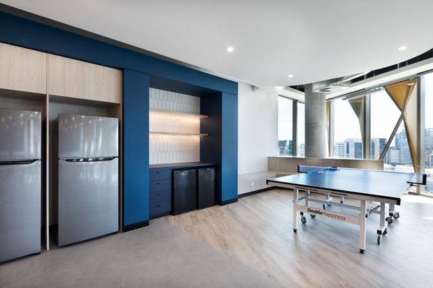 Bright contemporary recreational lounge with a full-size table tennis table, multiple refrigerators, built-in dark blue cabinetry, open shelving, and large floor-to-ceiling windows overlooking a city. Light timber flooring and exposed structural elements create an airy, modern communal space.