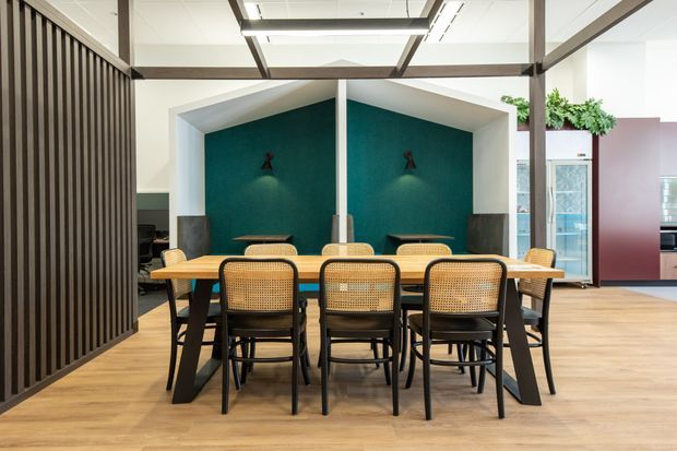 A contemporary communal dining and meeting area with a long timber table surrounded by black-framed woven cane chairs. The space features warm timber flooring, a deep teal geometric feature wall with wall lighting, exposed dark structural beams, and a black vertical-slat partition. Glazed doors and indoor greenery are visible in the background.