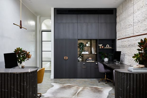 A refined modern home office with a dark built-in storage wall, integrated desk, open shelving, and concealed cabinetry. The room features a textured pale stone wall, polished concrete-style flooring, sculptural dark timber consoles, a marble-topped sideboard, a cowhide rug, and contemporary pendant lighting.