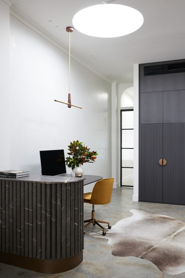 A refined home office with a sculptural dark fluted reception-style desk, marble-look benchtop, black monitor, and mustard leather task chair. The room features white walls with classic cornice detailing, polished concrete flooring, a large circular pendant light, and a full-height dark timber built-in storage wall with integrated ventilation and a concealed door. A neutral cowhide rug adds texture.