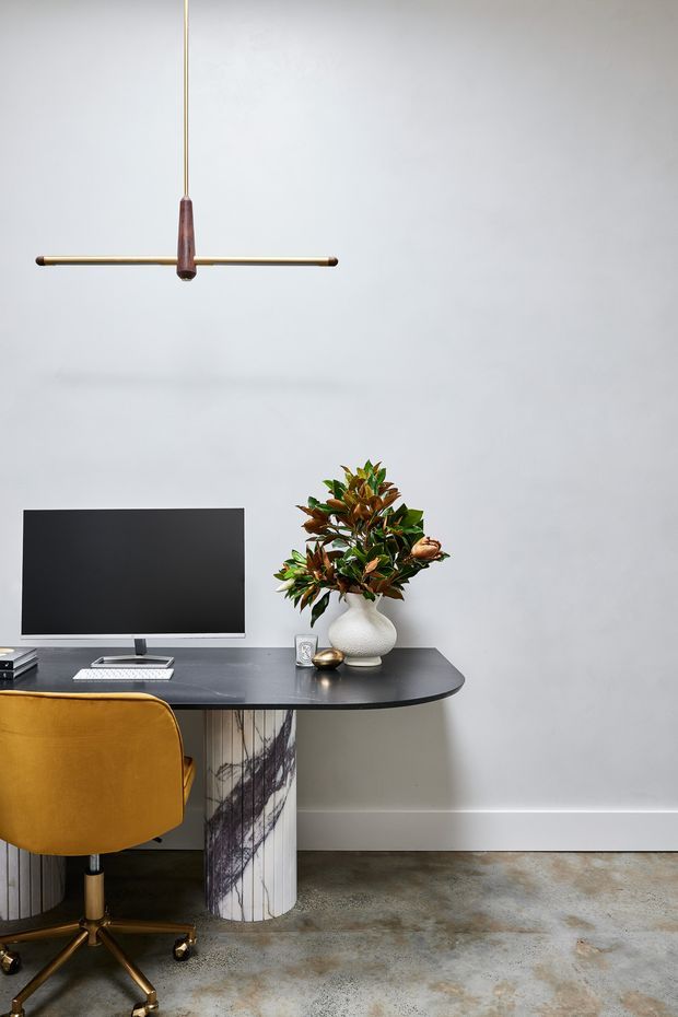 A refined compact home office with a black rounded-edge desk, computer monitor, mustard upholstered office chair, decorative vase and plant, and a minimalist linear pendant light. The desk has distinctive cylindrical marble-patterned supports, while the room features pale grey walls, a white skirting board, and a polished concrete floor.