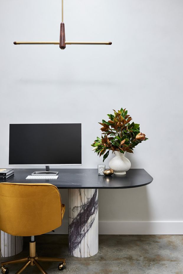 A compact, styled home office with a wall-mounted black monitor on a dark rounded-edge desk, a mustard upholstered swivel chair, decorative cylindrical desk supports, a sculptural pendant light, and a leafy arrangement in a textured white vase. The room has pale grey walls, a polished concrete floor, and a refined contemporary aesthetic with warm timber and brass accents.