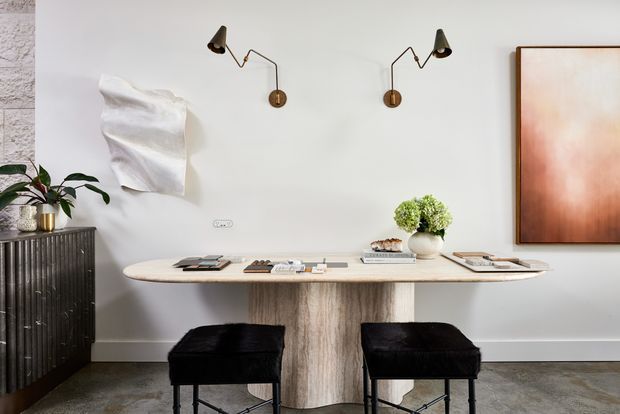 A refined dining or work nook with a sculptural light stone table, two black upholstered stools, wall-mounted brass swing-arm sconces, and a dark fluted cabinet topped with a plant. The room has pale walls, a polished concrete-like floor, abstract artwork, and restrained contemporary styling.