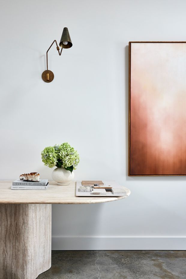 A refined minimalist interior vignette featuring a pale stone or travertine console table with a rounded edge and pedestal base, styled with books, magazines, a vase of green hydrangeas, and a small decorative object. A slim bronze wall sconce and a large framed abstract artwork add warmth against a light grey wall. The floor appears to be polished concrete, creating a calm, contemporary atmosphere.