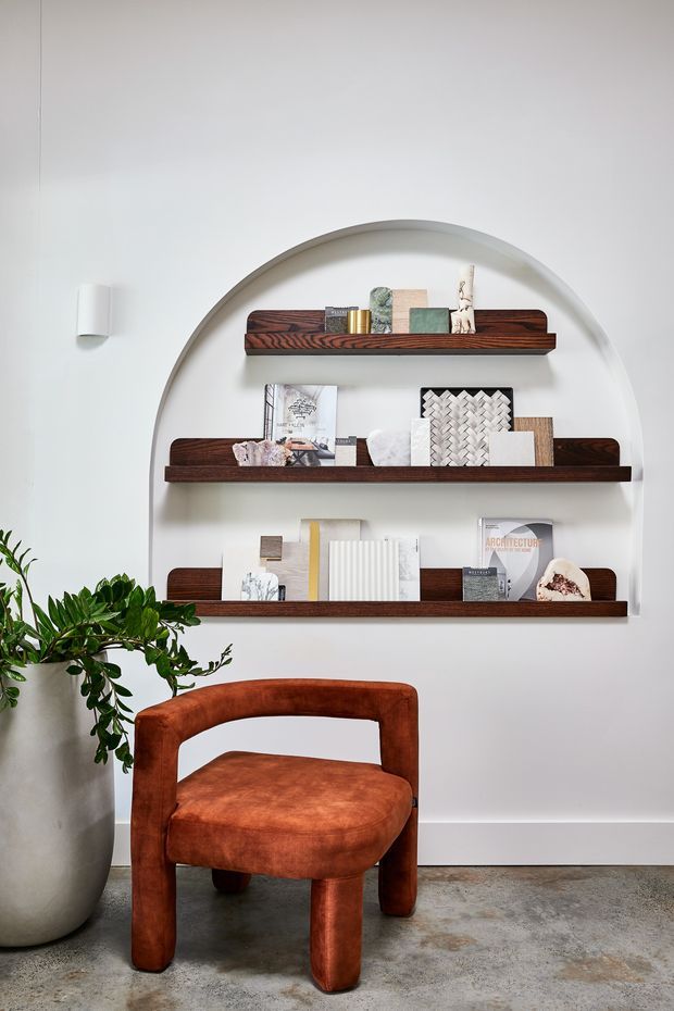 A refined interior vignette featuring a sculptural arched wall niche with three dark timber display shelves, styled with books, artwork, samples, and decorative objects. A rust-brown upholstered accent chair sits below, beside a large potted plant. The space has white walls, a concrete-look floor, and understated contemporary detailing.