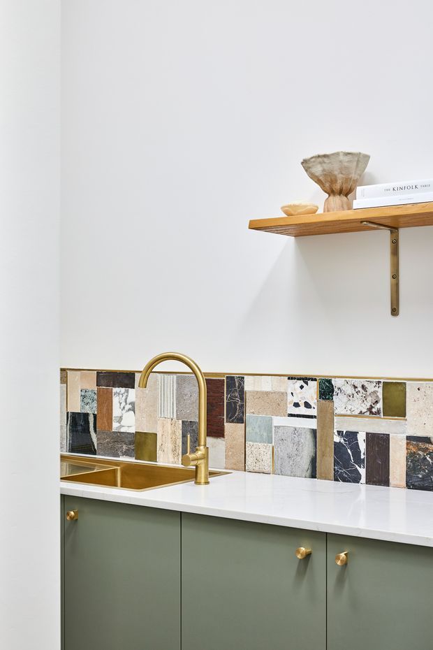 A bright, compact kitchen featuring muted sage-green cabinetry with brass knobs, a white benchtop, and a brass gooseneck tap over a brass-toned sink. The backsplash is a colourful patchwork of varied stone and tile pieces. Above, a simple timber shelf with brass brackets displays neutral-toned decorative objects and books. The overall look is refined, eclectic, and warm against white walls.