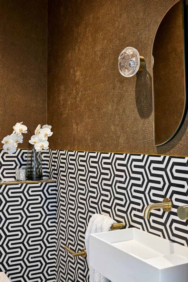 A dramatic, highly stylised bathroom vanity area featuring bold black-and-white geometric patterned wall tiles, a white rectangular vessel basin, brushed brass wall-mounted tapware, a textured bronze-brown wall finish, curved mirror, crystal wall sconce, and orchid arrangement.