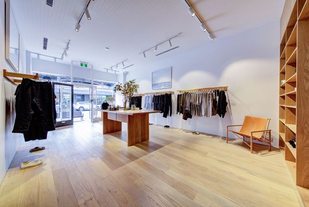Bright contemporary clothing boutique with light timber flooring, white paneled ceiling, track lighting, wall-mounted clothing rails, a central timber display table, timber shelving, and a glass storefront. A mid-century-style lounge chair and indoor plant add warmth to the minimalist retail interior.