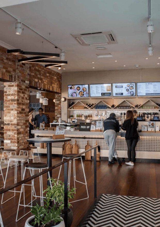 Contemporary café or casual food-and-drink venue interior with a long service counter, illuminated menu boards, exposed brick walls, bottle displays, patterned counter front, dark timber flooring, pendant and recessed lighting, and high tables with stools. Customers and staff are present, with a small planted area and bold black-and-white zigzag rug near the entrance.