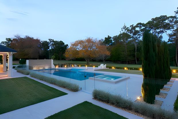 A landscaped outdoor pool area at dusk, featuring a rectangular swimming pool with a raised spa, transparent glass safety fencing, pale paved paths, manicured lawn, ornamental grasses, sculpted trees, and warm recessed landscape lighting. A pavilion or building edge is visible at the left.