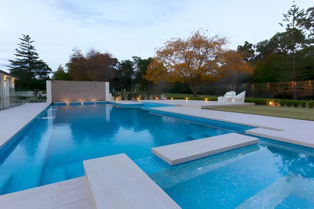 A landscaped residential outdoor pool area at dusk, featuring a large modern geometric swimming pool with integrated shallow sections and broad pale paving. The pool is surrounded by lawns, mature trees, outdoor lighting, and contemporary buildings, creating a refined resort-like setting.