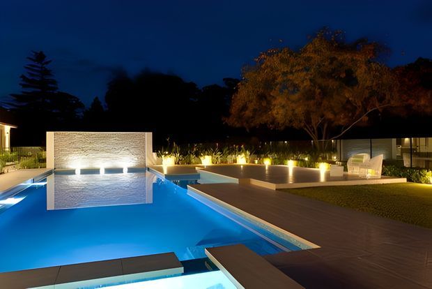 Night-time view of a contemporary landscaped pool area with a large illuminated swimming pool, raised water feature with textured stone wall, broad timber-toned decking, integrated low seating or retaining walls, and warm architectural landscape lighting. Mature trees and planted borders frame the outdoor entertaining space.
