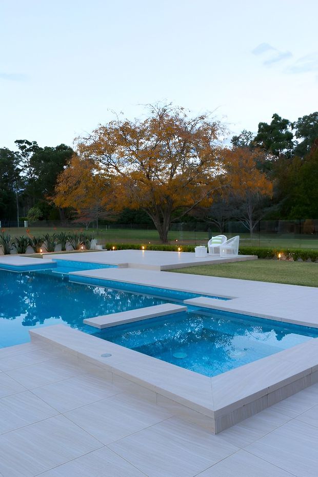 A landscaped outdoor pool area with a large swimming pool and raised square spa, surrounded by pale tiled paving, a manicured lawn, garden planting, and outdoor seating. The scene is framed by mature trees with autumn foliage and appears calm and neatly designed.