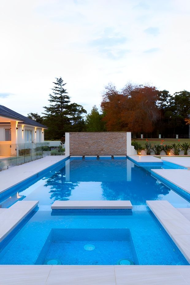 A modern outdoor swimming pool complex at dusk, featuring a large vivid-blue tiled pool with shallow entry areas, raised rectangular platforms, broad pale paving, glass balustrades, planted garden beds, and a stone-clad feature wall. A contemporary single-storey house is visible to the left, with mature trees forming the background.